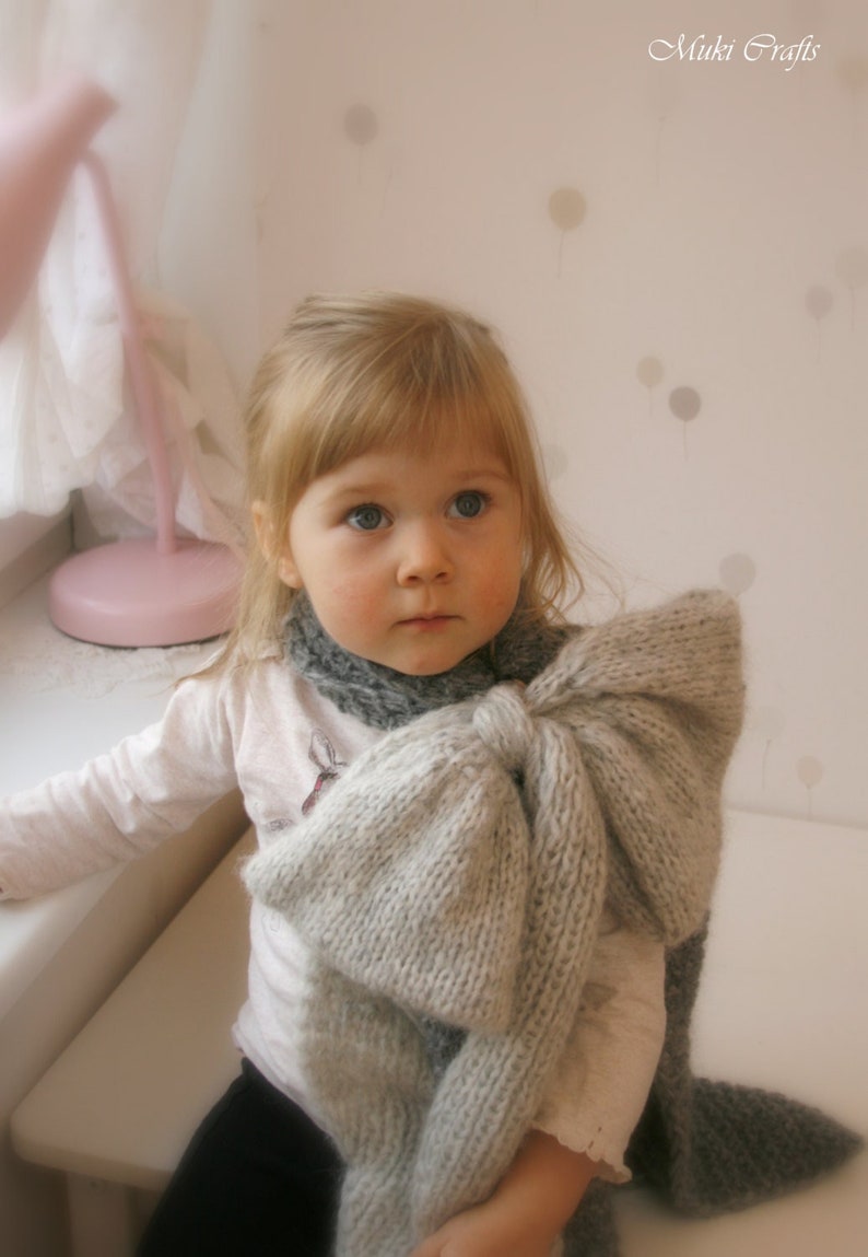 May include: A young girl wearing a white shirt and black pants with a large, gray knitted bow tied around her neck. The bow is made of a soft, fluffy yarn and is tied in a large, dramatic knot.