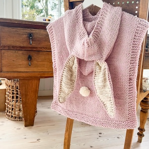 May include: A light pink knitted poncho with a hood and bunny ears. The ears are beige and attached to the front of the poncho, along with a small white pom-pom. The poncho is hanging on a wooden chair.