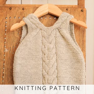 May include: A light grey knitted baby vest with a cable knit design down the centre, hanging on a wooden hanger. The text "KNITTING PATTERN" is displayed at the bottom of the image. The vest is made of a soft, textured yarn.