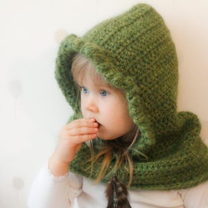 Peut inclure: Une capuche verte au crochet avec une plume. La capuche est d'un vert vif et semble faite &agrave; la main. L'enfant porte un haut blanc &agrave; manches longues. Le fond est blanc doux avec des formes claires subtiles.