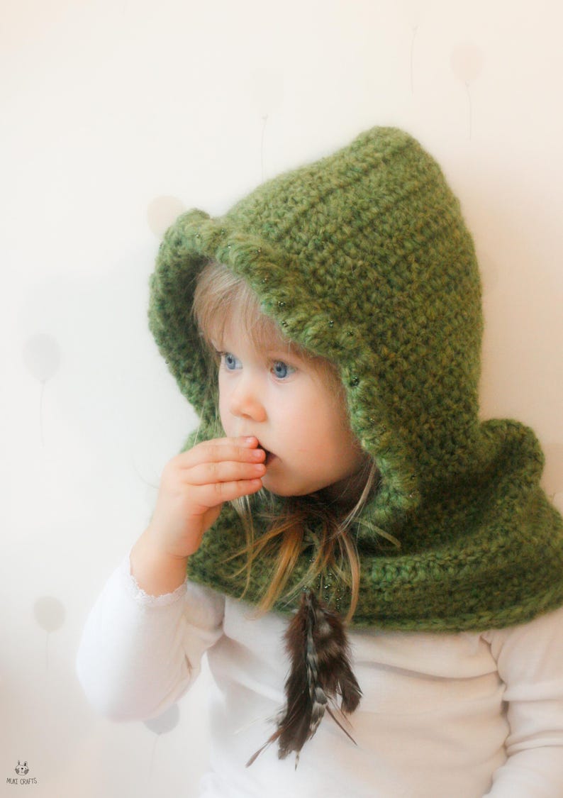Peut inclure: Une capuche verte au crochet avec une plume. La capuche est d'un vert vif et semble faite &agrave; la main. L'enfant porte un haut blanc &agrave; manches longues. Le fond est blanc doux avec des formes claires subtiles.