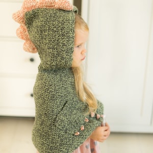 Peut inclure: Un sweat &agrave; capuche dinosaure crochet&eacute; vert et p&ecirc;che avec une capuche et des pointes. Le sweat &agrave; capuche est port&eacute; par un enfant, et les manches ont des accents de couleur p&ecirc;che. L'enfant porte une jupe rose avec des c&oelig;urs gris. Le logo Muki Crafts est dans le coin sup&eacute;rieur droit.
