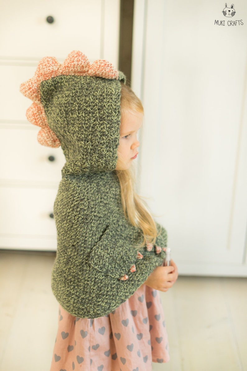 Peut inclure: Un sweat &agrave; capuche dinosaure crochet&eacute; vert et p&ecirc;che avec une capuche et des pointes. Le sweat &agrave; capuche est port&eacute; par un enfant, et les manches ont des accents de couleur p&ecirc;che. L'enfant porte une jupe rose avec des c&oelig;urs gris. Le logo Muki Crafts est dans le coin sup&eacute;rieur droit.