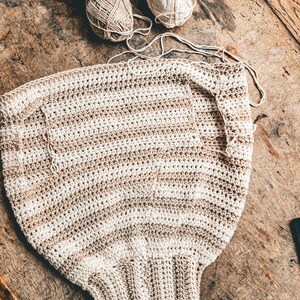 May include: A partially completed crochet project in cream and tan stripes, with several balls of yarn in similar colors scattered around. The project appears to be a garment, possibly a top or sweater, and is laid out on a wooden surface.
