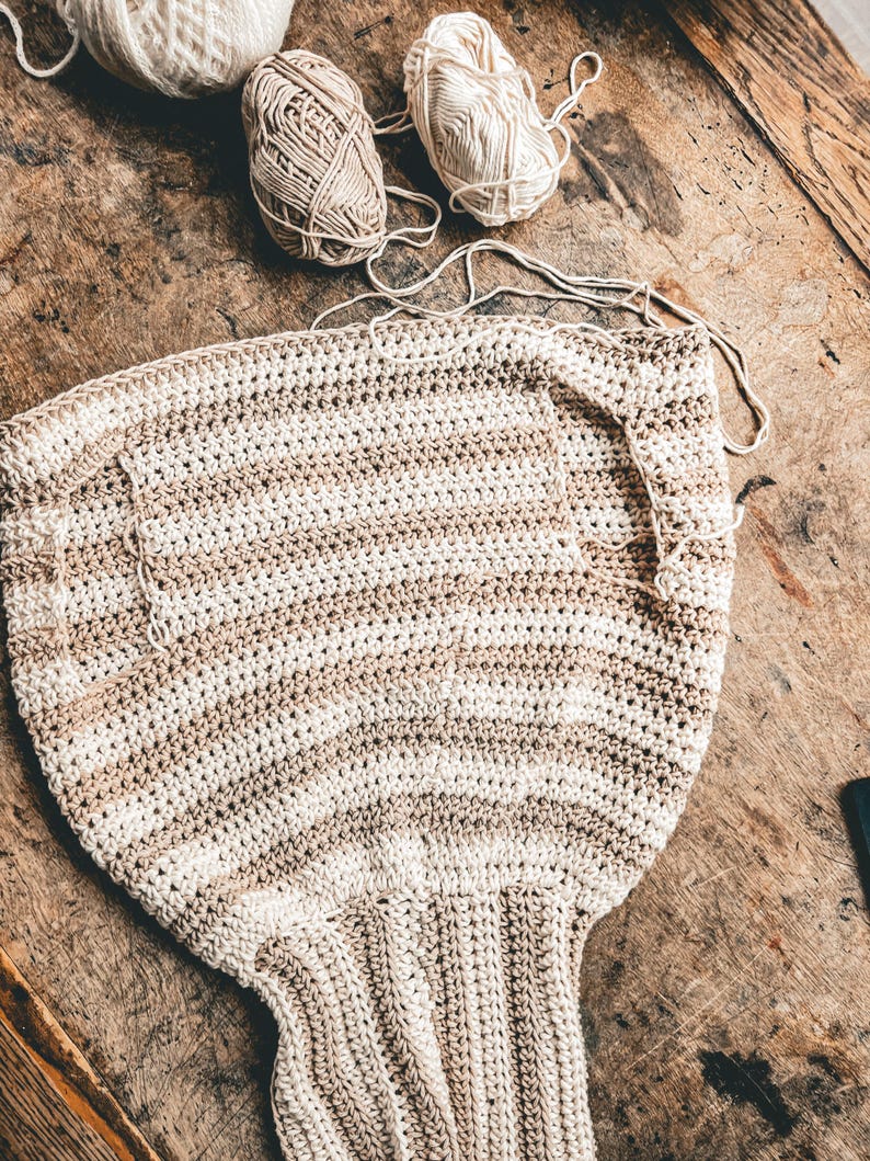 May include: A partially completed crochet project in cream and tan stripes, with several balls of yarn in similar colors scattered around. The project appears to be a garment, possibly a top or sweater, and is laid out on a wooden surface.