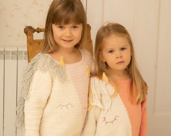 May include: Two long-sleeved sweaters featuring a unicorn design with a cream and pastel pink or coral color block pattern. Each sweater includes a yellow horn, a fringe mane, and embroidered closed eyes. The text KNITTING PATTERN is visible.