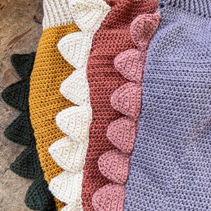 May include: A collection of four crocheted garments featuring ribbed necklines and rows of triangular spikes along the side. The items are colored in dark green, mustard yellow, cream, terracotta, and lavender, each with matching spikes.