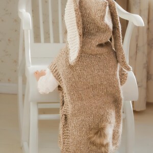 May include: A knitted bunny costume in a light brown color, featuring long ears with cream-colored inner lining and a fluffy tail. The costume is worn by a child, standing near a white rocking chair. The outfit is handmade.