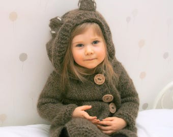 May include: A child wearing a brown knitted bear-themed hooded coat with wooden buttons. The coat has bear ears on the hood. The text "KNITTING PATTERN" is displayed at the bottom of the image.