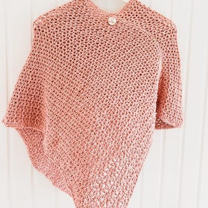 May include: A light pink crocheted poncho with a button closure. The poncho is hanging on a white hanger against a white background. The crochet pattern is visible, and the garment appears soft and textured.