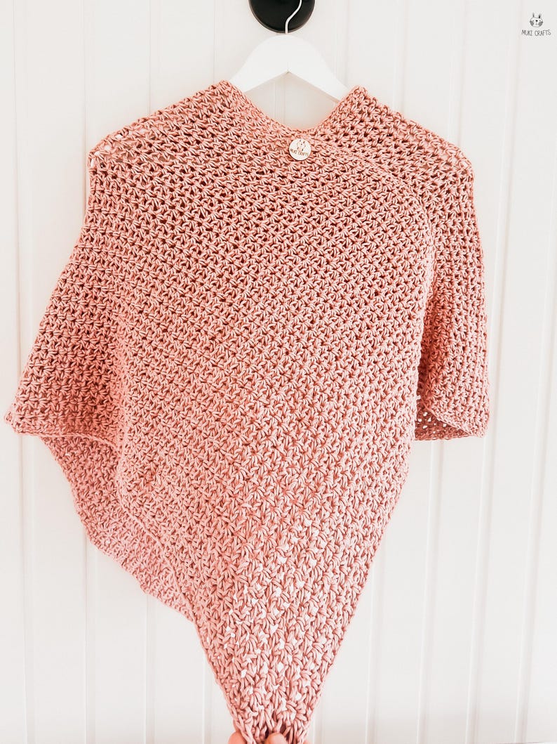 May include: A light pink crocheted poncho with a button closure. The poncho is hanging on a white hanger against a white background. The crochet pattern is visible, and the garment appears soft and textured.