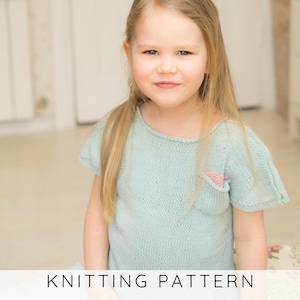 May include: A light blue knitted top with short sleeves and a small pink pocket. The text "KNITTING PATTERN" is displayed at the bottom of the image. The top is worn by a young child with long blonde hair.