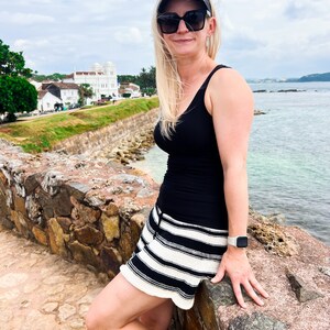 May include: A woman wearing a black tank top, black and white striped shorts, and a visor. She is wearing sunglasses and a smartwatch. The background features a stone wall, ocean, and buildings under a cloudy sky.