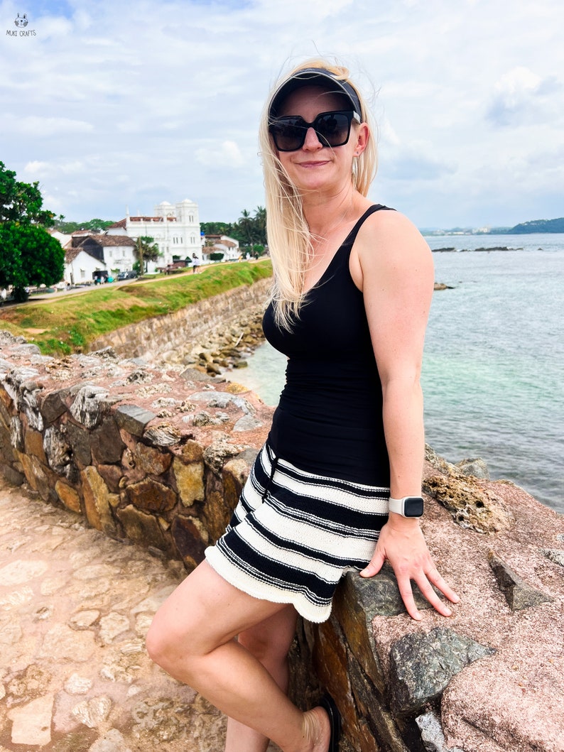 May include: A woman wearing a black tank top, black and white striped shorts, and a visor. She is wearing sunglasses and a smartwatch. The background features a stone wall, ocean, and buildings under a cloudy sky.