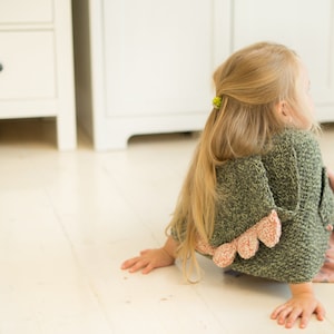 Peut inclure: Un cardigan vert tricot&eacute; pour enfant avec une bordure festonn&eacute;e rose. Le cardigan fait partie d'une collection de v&ecirc;tements pour enfants. L'enfant est sur un sol en bois clair, avec de longs cheveux blonds et un chouchou jaune.
