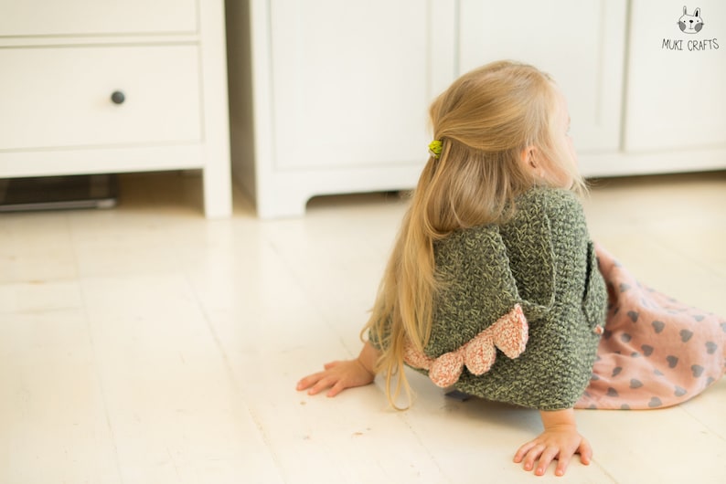 Peut inclure: Un cardigan vert tricot&eacute; pour enfant avec une bordure festonn&eacute;e rose. Le cardigan fait partie d'une collection de v&ecirc;tements pour enfants. L'enfant est sur un sol en bois clair, avec de longs cheveux blonds et un chouchou jaune.