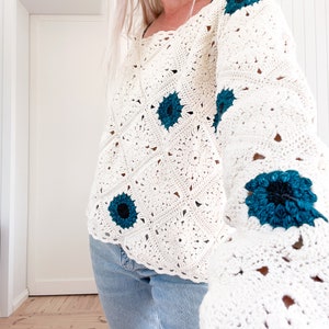 May include: Cream-colored crochet sweater with teal flower accents. The sweater features a square neckline and long sleeves. The floral details are centered with a black dot. The sweater is paired with blue jeans.