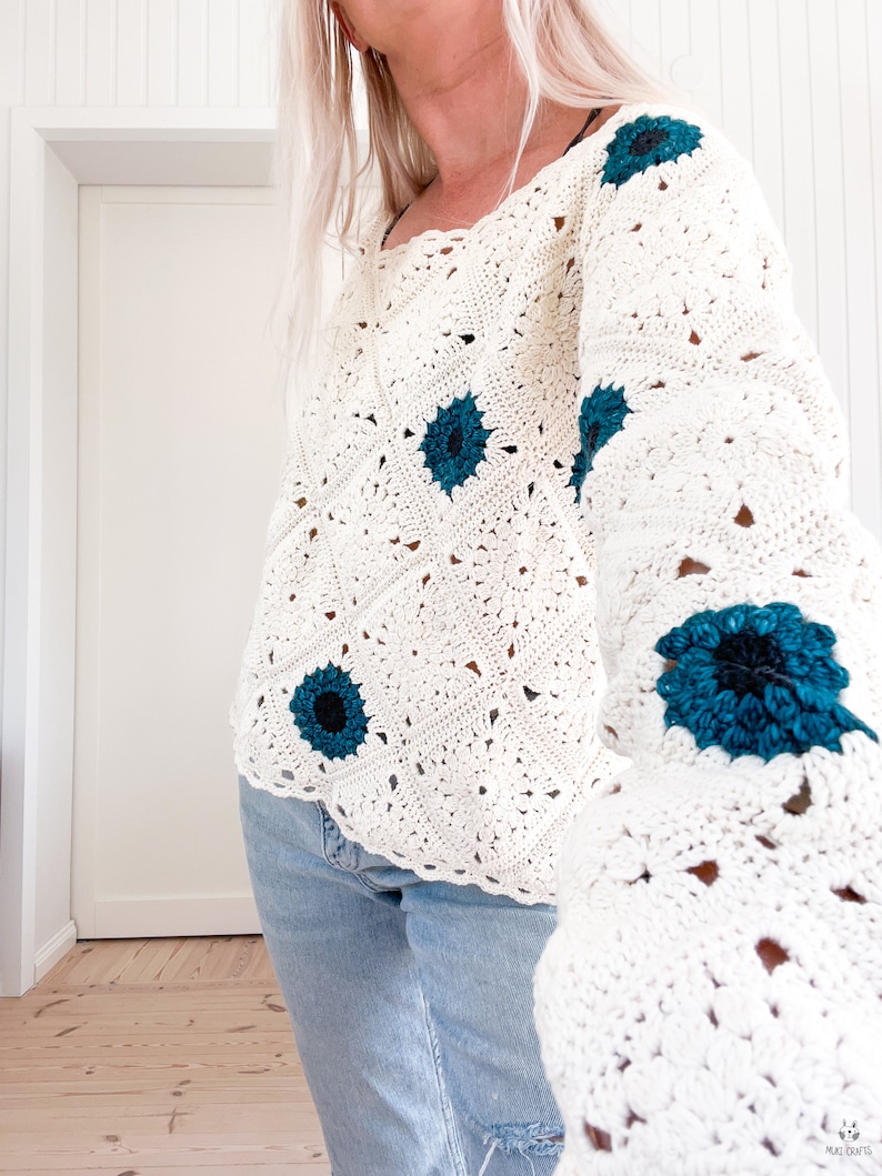 May include: Cream-colored crochet sweater with teal flower accents. The sweater features a square neckline and long sleeves. The floral details are centered with a black dot. The sweater is paired with blue jeans.