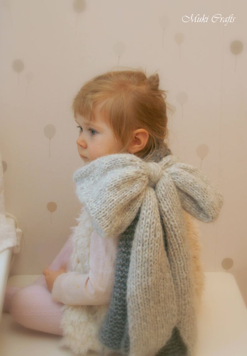 May include: A young child wearing a white knitted vest and a large, gray knitted bow tied around their neck. The bow is tied in a large, loose knot and hangs down the child's back.