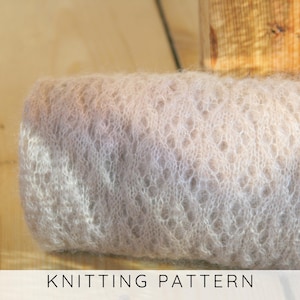 Op de afbeelding: Een opgerold stuk lichtbeige, pluizige gebreide stof in mohair-stijl met een delicaat, opengewerkt ruitpatroon. De textuur is zacht en luchtig, met zichtbare vezels die een halo-effect creëren over het oppervlak van het gebreide materiaal.