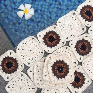May include: Crocheted square motifs in cream, brown, and black are arranged on a gray surface. Each square features a central brown and black design, surrounded by a cream-colored border. A white flower floats in a blue tiled pool.