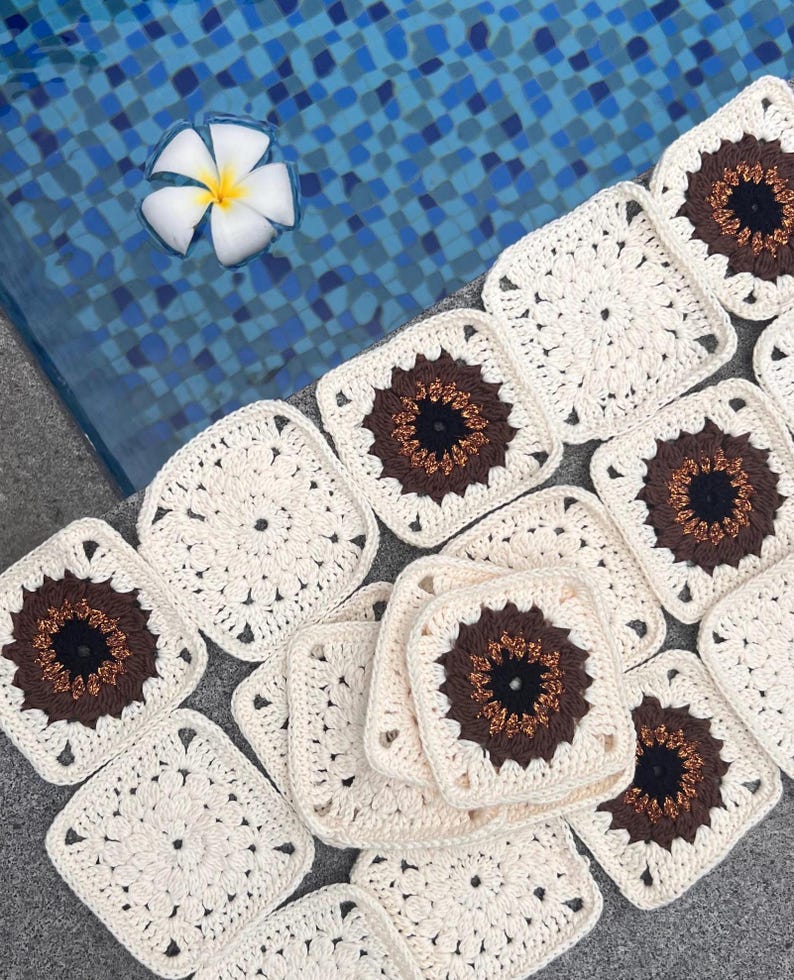 May include: Crocheted square motifs in cream, brown, and black are arranged on a gray surface. Each square features a central brown and black design, surrounded by a cream-colored border. A white flower floats in a blue tiled pool.
