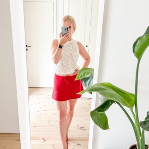 May include: A sleeveless, cream-colored lace top paired with a bright red mini skirt and matching red heels. The outfit is captured in a mirror, reflecting a modern, stylish ensemble. The image showcases a fashion-forward look.