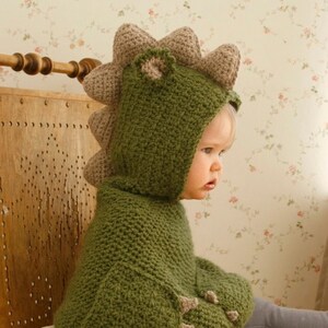 Peut inclure: Un sweat &agrave; capuche dinosaure vert crochet&eacute; avec une capuche et des &eacute;pines attach&eacute;es. Le sweat &agrave; capuche a des d&eacute;tails en forme de griffes sur les manches. Le texte "Muki Crafts" est visible. L'enfant porte un pantalon gris.