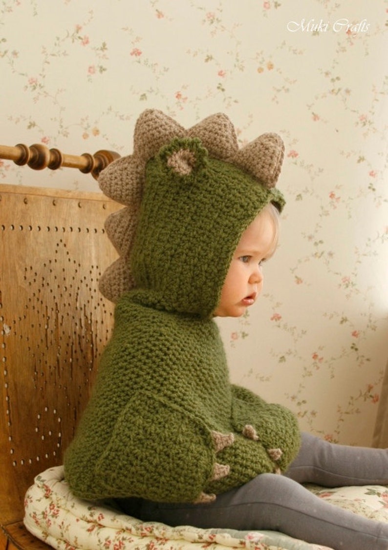 Peut inclure: Un sweat &agrave; capuche dinosaure vert crochet&eacute; avec une capuche et des &eacute;pines attach&eacute;es. Le sweat &agrave; capuche a des d&eacute;tails en forme de griffes sur les manches. Le texte "Muki Crafts" est visible. L'enfant porte un pantalon gris.