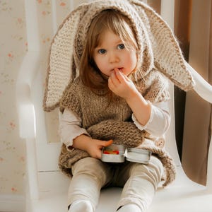 May include: A young child wearing a knitted bunny ear hood and vest, sitting on a white chair. The child is holding a small tin with colorful candies. The outfit is a light brown color, and the child is wearing beige pants and white socks.