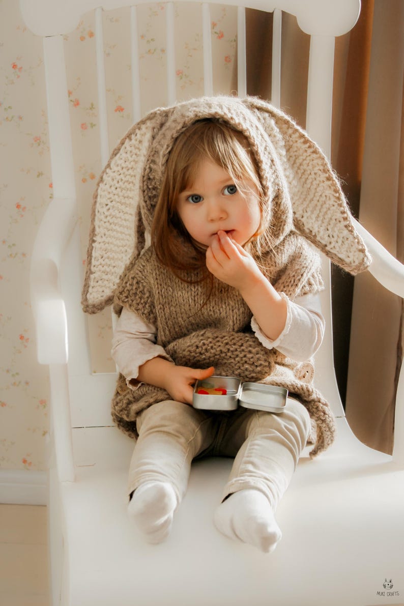 May include: A young child wearing a knitted bunny ear hood and vest, sitting on a white chair. The child is holding a small tin with colorful candies. The outfit is a light brown color, and the child is wearing beige pants and white socks.