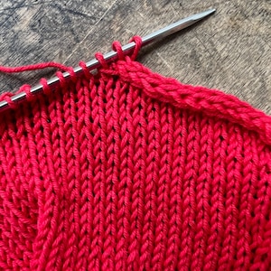May include: Close-up of red knitted fabric on a wooden surface. A silver knitting needle holds several loops of yarn, with the knitted rows forming a textured pattern. The vibrant red color of the yarn contrasts with the wood grain.