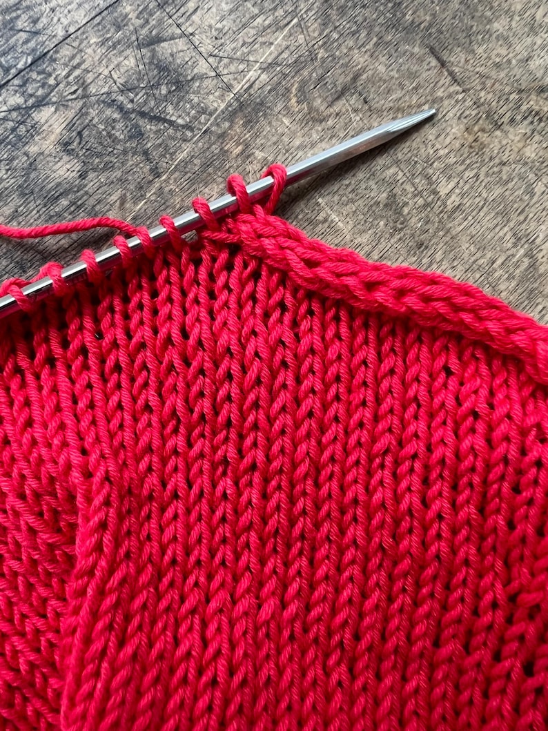 May include: Close-up of red knitted fabric on a wooden surface. A silver knitting needle holds several loops of yarn, with the knitted rows forming a textured pattern. The vibrant red color of the yarn contrasts with the wood grain.