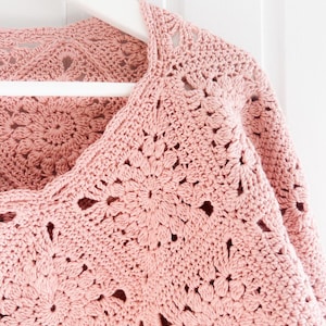 May include: A light pink, open-weave crochet top with a floral pattern. The top is displayed on a white hanger against a white background. The crochet design features a repeating flower motif, creating a delicate and textured appearance.