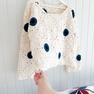 Op de afbeelding: Cr&egrave;mekleurige gehaakte trui met een vierkante halslijn en lange mouwen. De trui heeft een ruitpatroon en is versierd met marineblauwe bloemenapplicaties. Het kledingstuk hangt aan een witte hanger tegen een witte muur.