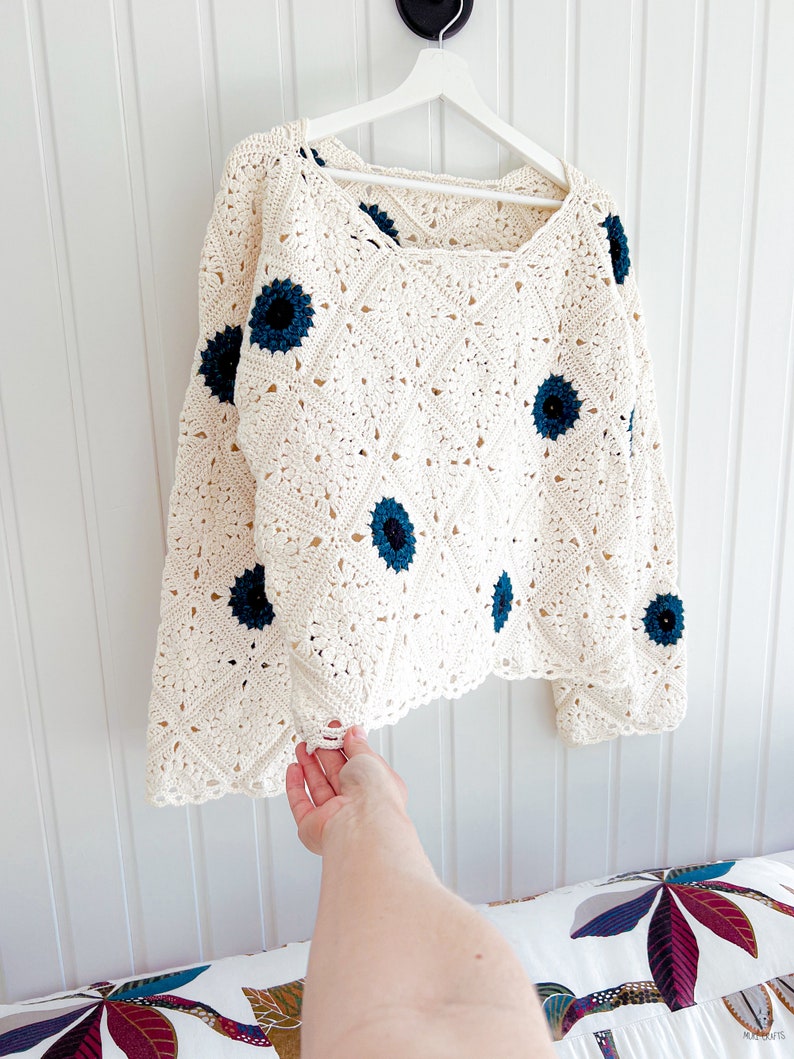Op de afbeelding: Cr&egrave;mekleurige gehaakte trui met een vierkante halslijn en lange mouwen. De trui heeft een ruitpatroon en is versierd met marineblauwe bloemenapplicaties. Het kledingstuk hangt aan een witte hanger tegen een witte muur.
