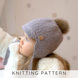 Pode incluir: Um gorro cinza tricotado com protetores de orelha e um pompom de pele marrom. O chapéu apresenta um botão de madeira e está rotulado "KNITTING PATTERN" em texto branco na parte inferior. Perfeito para o inverno.