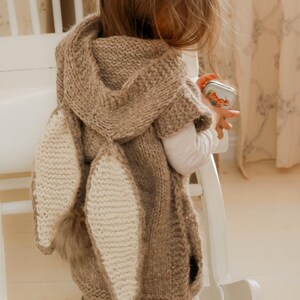 May include: A brown knitted hooded cape with large, cream-colored bunny ears and a fluffy tail. The cape is worn by a child, and the design is reminiscent of a rabbit. The cape is a cozy and whimsical garment.