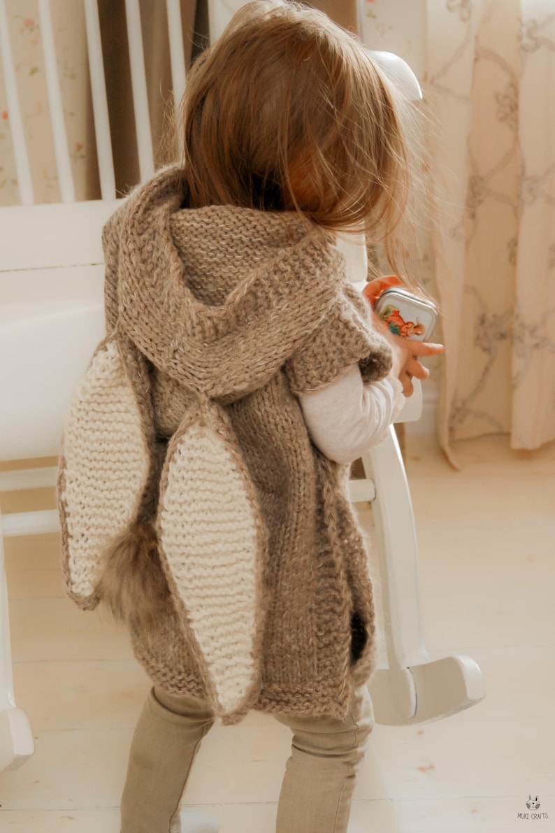 May include: A brown knitted hooded cape with large, cream-colored bunny ears and a fluffy tail. The cape is worn by a child, and the design is reminiscent of a rabbit. The cape is a cozy and whimsical garment.