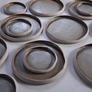 May include: A collection of ceramic plates in various sizes and shapes, all with a gray glaze and a slightly irregular rim. The plates are arranged on a white surface.