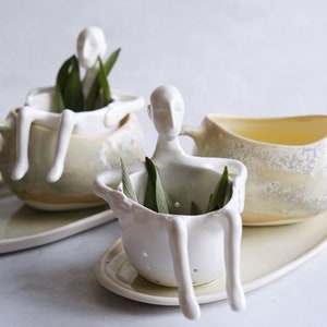 May include: Two white ceramic teacups with small human figures holding sprigs of green herbs. The teacups are on matching oval-shaped saucers. One teacup has a strainer.