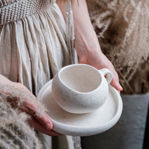 May include: A speckled white ceramic teacup and saucer set. The cup is round with a small handle, and the saucer is a matching flat circle. The set is held in front of a beige dress and dried plants.