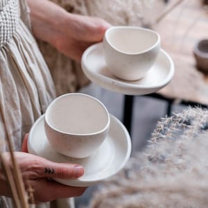 May include: Two off-white ceramic teacups and saucers are held. The cups have a rounded shape and speckled texture. The saucers are round and flat. The background includes additional ceramic pieces and dried plants, suggesting a rustic or minimalist aesthetic.