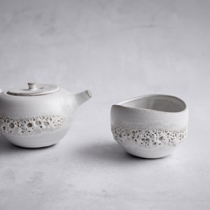 Puede incluir: Una tetera y una taza de té de cerámica blanca con un diseño texturizado con forma de burbujas.