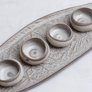 May include: A handmade ceramic serving tray with four small bowls. The tray is oblong with a textured, crackled surface. The bowls are round with a textured exterior and a smooth, white interior. The rims of the bowls are a darker shade of brown.