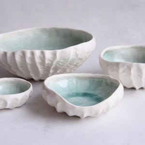 Pode incluir: Conjunto de quatro tigelas de cerâmica branca com um design texturizado em forma de concha. As tigelas são envidraçadas em uma cor azul clara.