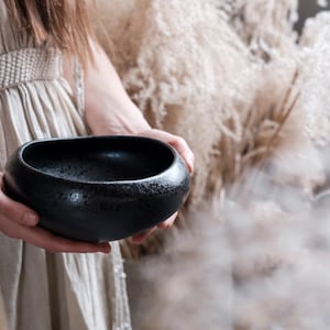 Puede incluir: Un cuenco de cerámica negra con textura moteada se sostiene con ambas manos. El cuenco tiene una forma redondeada y orgánica y un acabado oscuro y mate. El fondo presenta tonos suaves y neutros y una estética natural borrosa.