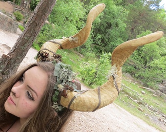 May include: A large, curved, tan-colored horn headpiece with brown straps and decorative greenery. The horns are attached to a headband adorned with moss and small pinecones. The headpiece is worn by a person with long brown hair.