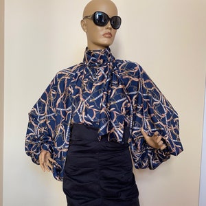 May include: A blue and gold patterned blouse with a high neck and long sleeves. The blouse has a gathered detail at the neck and a loose, flowing fit. The sleeves are gathered at the shoulders and have a wide, bell-shaped cuff.