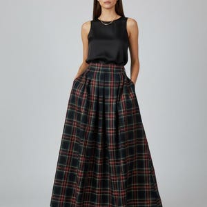 Puede incluir: Una mujer con una blusa negra sin mangas y una falda larga plisada a cuadros. La falda presenta un patrón de líneas verdes oscuras, rojas y amarillas. El atuendo se completa con zapatos beige y un collar dorado.